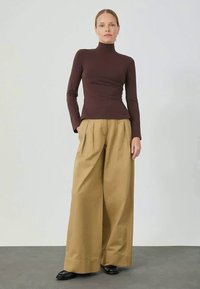 Rjava turtleneck majica v kombinaciji z visokim pasom in širokimi khaki hlačami. Črni čevlji, enostavna ozadja, gladke teksture in nevtralni toni.