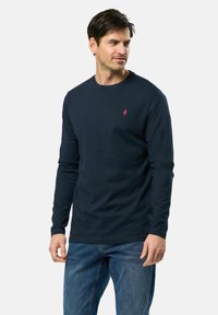 Chemise à manches longues marine en coton, avec un col rond et un petit logo rouge sur la poitrine. Associée à un jean en denim bleu.