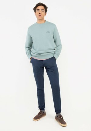 Joven de pie con las manos en los bolsillos, lleva una sudadera verde claro, pantalones azul marino, calcetines grises y zapatillas marrones sobre fondo blanco.