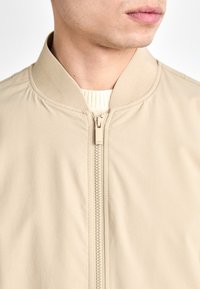 Beige bomberjakke med en glatt tekstur, ribbet krage og glidelåsdetaljer foran. Subtil merkevare nær glidelåsen.