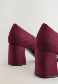 Zapatos de tacón alto de ante burdeos con tacones gruesos. El diseño presenta una punta afilada y una textura suave con una suela negra brillante.
