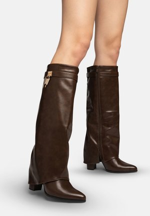 Nogi w ciemnobrązowych skórzanych botkach za kolano z niskim obcasem typu blok i złotymi suwakami na neutralnym tle.