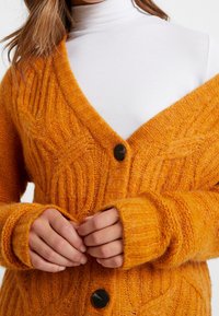 Cardigan arancione con trama, realizzato in materiale morbido, caratterizzato da una scollatura a V profonda, un motivo a costine e una chiusura con un singolo bottone nero.