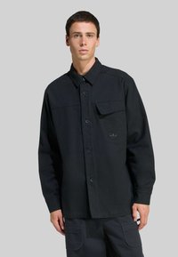 Mladý muž oblečený v čiernej košeli na gombíky s dlhými rukávmi a náprsnou kapsou, skombinovanej s tmavými cargo nohavicami na šedom pozadí.