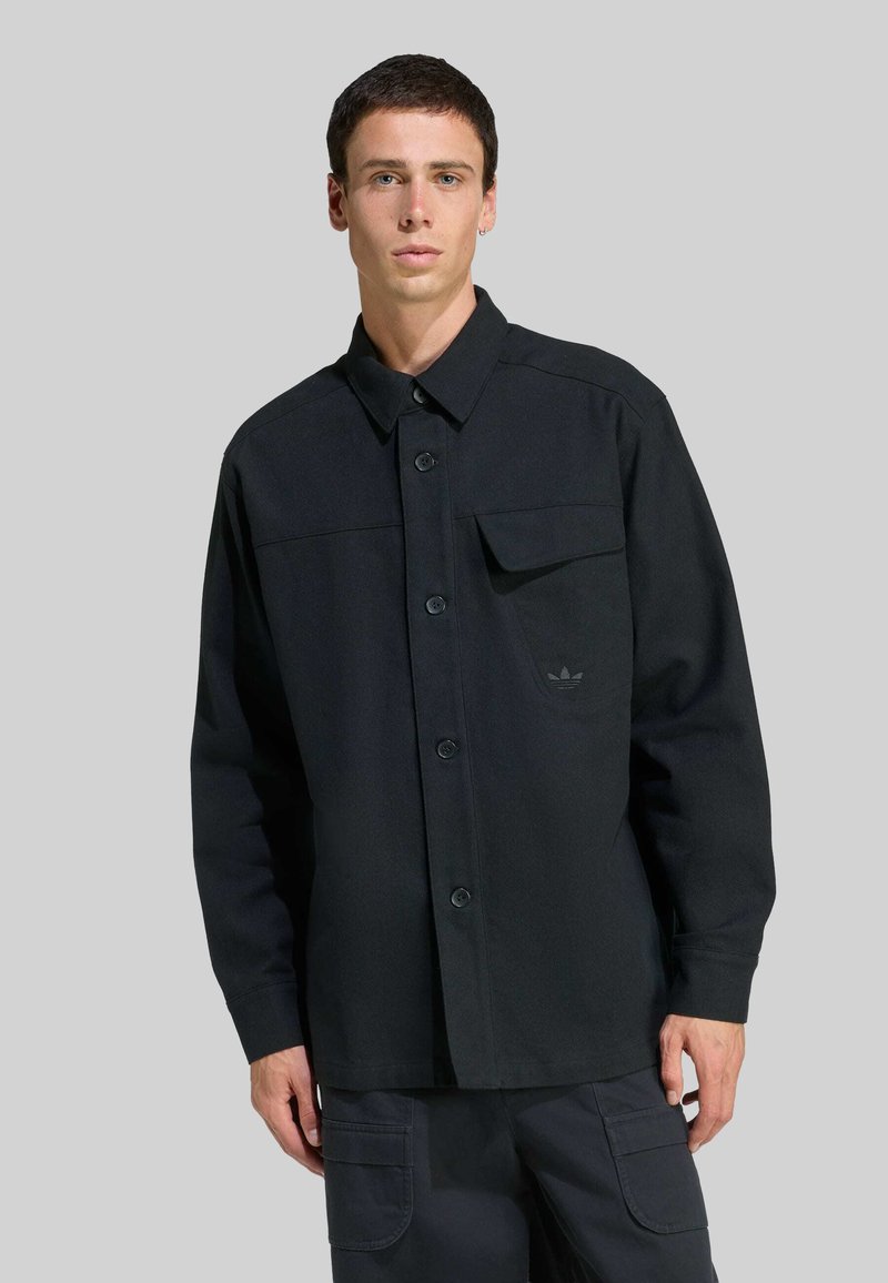 Mladý muž oblečený v čiernej košeli na gombíky s dlhými rukávmi a náprsnou kapsou, skombinovanej s tmavými cargo nohavicami na šedom pozadí.