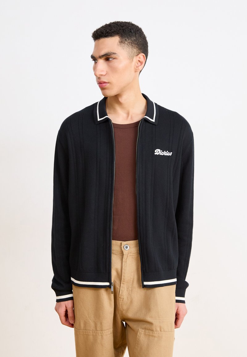 Cardigan nero con zip, texture a coste, colletto e orlo bianchi a contrasto, con logo ricamato sul petto. Indossato sopra una camicia marrone.