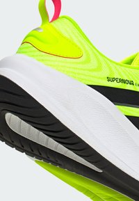 Neon-gelber und schwarzer Sportschuh mit dicker weißer Sohle, roten und gelben Zugschlaufen, strukturierter Sohle und "SUPERNOVA"-Logo an der Seite.
