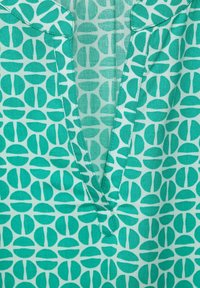 Zielony top w wzory wykonany z lekkiej tkaniny, charakteryzujący się abstrakcyjnym wzorem półokręgów. Wyraźny dekolt w serek oraz jeden detal kieszeni.