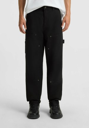 Man wearing black loose-fit pants with multiple pockets and black sneakers, standing against a plain light background.