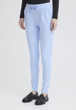 Žena v svetlomodrých úzkych nohaviciach so šnúrkou v páse a čiernych mokasínach, stojaca pred jednoduchým svetlým pozadím.