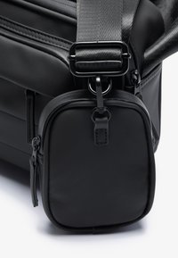 Borsa sintetica nera con manico testurizzato, dotata di una piccola tasca staccabile, chiusura con zip e delicati dettagli del logo sul lato.