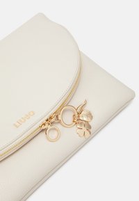 Crèmekleurige clutch van nepleer met gouden rits, met decoratieve bedelaccenten waaronder een lieveheersbeestje en klavertjevier. Merkje namens in goud geperst.
