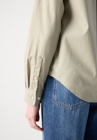 Beige long-sleeve shirt with a smooth texture, featuring buttoned cuffs and a curved hem, paired with blue denim jeans.