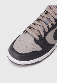 Sneaker z szarym skórzanym wierzchem z perforowanym czubkiem, ciemnoszarymi akcentami i jasnoszarymi sznurowadłami. Biała podeszwa środkowa z teksturowaną czarną podeszwą.