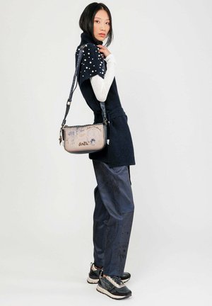 Frau in navyblauen Hosen, schwarzem Kurzarmpullover mit Perlendetails, weißem Langarmshirt, schwarzen Sneakers und einer beigen Umhängetasche mit Illustrationen.