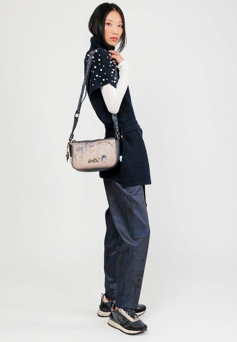 Donna con pantaloni blu navy, maglietta nera a maniche corte con dettagli in perle, camicia bianca a maniche lunghe, sneakers nere e borsa a tracolla beige con illustrazioni.