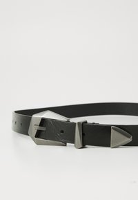 Ceinture en cuir noir avec une boucle triangulaire en argent mat et des accents pointus. Elle possède cinq trous réglables le long de la sangle pour ajuster la taille.