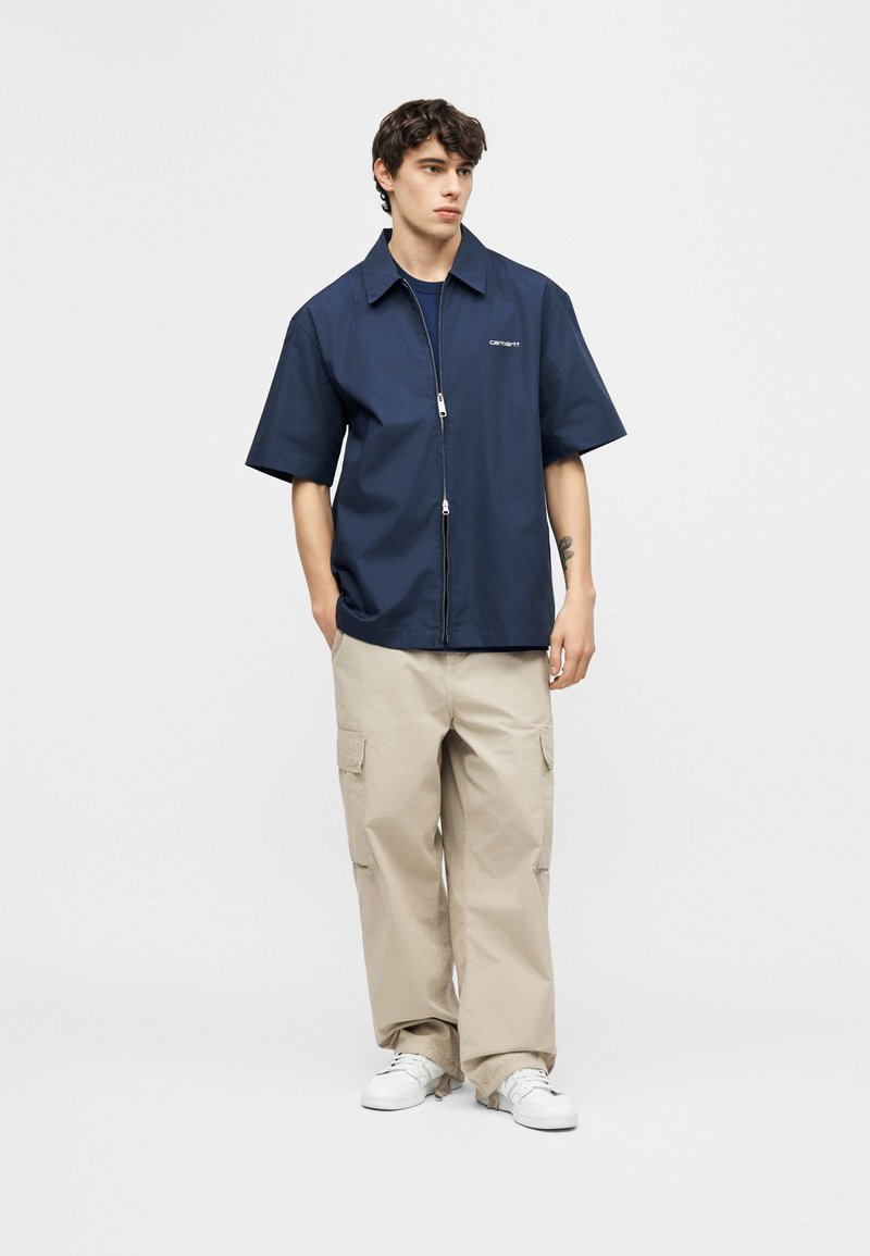 Jeune homme debout les mains dans les poches, portant une chemise zippée marine à manches courtes, un pantalon cargo beige et des baskets blanches.