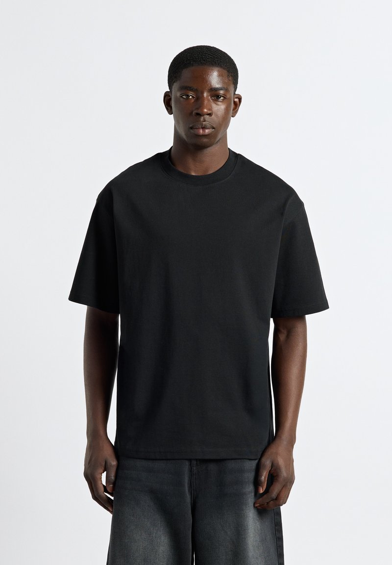 Junger Mann mit kurzen Haaren stehend, trägt ein schlichtes schwarzes übergroßes T-Shirt und dunkle Jeans vor einem einfarbigen weißen Hintergrund.