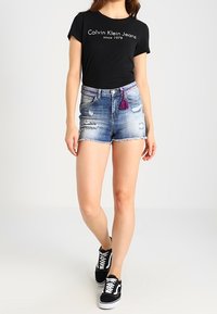 Czarny t-shirt z białym logo, podniszczone szorty dżinsowe z niebieskimi i fioletowymi akcentami oraz czarne trampki z białą podeszwą.