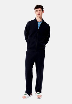 Trening din fleece de culoare albastră închisă, cu jachetă cu fermoar și pantaloni, asortați cu papuci albi cu un logo verde. Textură fină, croială lejeră.