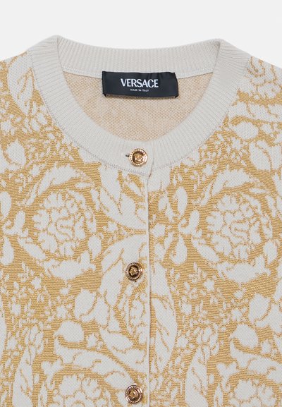 Cardigan lavorato a maglia color crema con motivo floreale dorato, dotato di un collo rotondo, polsini a coste e tre bottoni dorati sulla parte anteriore.
