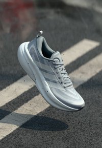 Chaussure de sport gris clair à texture lisse, avec trois bandes gris foncé, un col rembourré et une silhouette élégante.