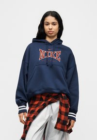 Μπλε σκούρο φούτερ Nike με τσέπη καγκουρό, κουκούλα με κορδόνι, λευκό και κόκκινο λογότυπο και ριγέ μανσέτες. Συνδυασμένο με κόκκινο και μαύρο πουκάμισο με σχέδιο καρό σε στρώσεις.