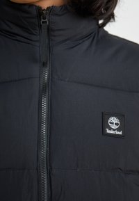 Czarna pikowana kamizelka z wysokim kołnierzem, pełnym zamkiem błyskawicznym oraz kwadratową naszywką z logo Timberland po lewej stronie. Gładka faktura i lekki materiał.