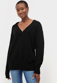 Sort striktrøje med dyb V-udskæring, dropped shoulders og ribbede manchetter. Blød tekstur, afslappet pasform, parret med blå jeans.