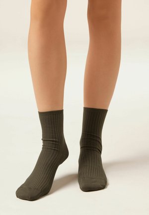 Beine, die dunkelolivgrüne gerippte Socken tragen, stehen auf einer hellen neutralen Oberfläche vor einem schlichten Hintergrund.