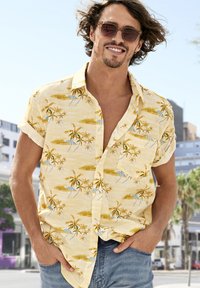 Maglietta gialla a maniche corte con stampa di palme e scena da spiaggia. Taschino frontale, tessuto leggero, vestibilità rilassata. Indossata con pantaloni in denim blu.