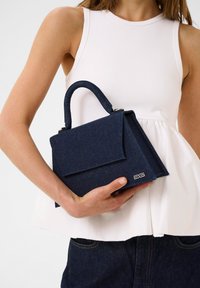Borsa in denim blu scuro con forma strutturata, manico superiore e chiusura a pattina. Include un dettaglio del logo sul lato.
