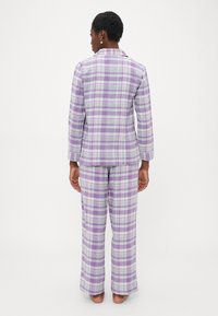 Žena stojaca naboso, pohľad zozadu, oblečená v fialovo-bielej kárované pyžamo s dlhými rukávmi a nohavicami.