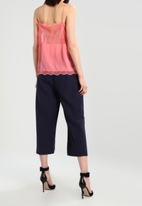 Camisole en dentelle rose et tissu transparent avec bretelles réglables, associée à un pantalon court bleu marine. Des chaussures à talons haut noires complètent la tenue.