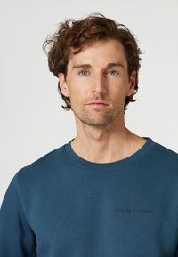 Sudadera de algodón azul con cuello redondeado. Presenta un sutil logo en relieve en la parte frontal. Textura suave, ajuste casual.