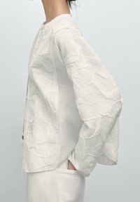 Donna che indossa una blusa bianca a maniche lunghe con texture e motivi floreali insieme a pantaloni bianchi, con le mani nelle tasche, mostrata di profilo contro uno sfondo semplice.