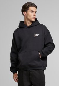Hoodie oversize nero in cotone con tasca a marsupio frontale e logo bianco sul petto sinistro. Polsini e orlo ribattuti.