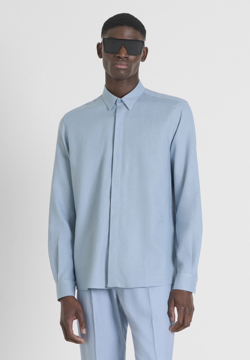 Camicia azzurro chiaro a maniche lunghe in un tessuto morbido con chiusura a bottoni, colletto e vestibilità slim. Il modello indossa occhiali da sole scuri in contrasto.