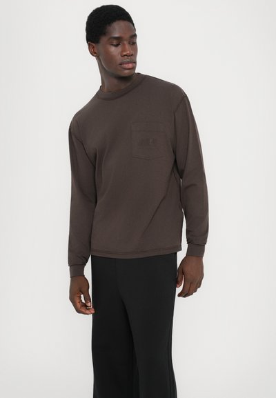 Brown long-sleeve t-shirt with a round neckline, featuring a chest pocket and subtle stitching details. Worn with black loose-fitting pants.
