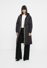 Black padded coat with a hood, featuring yellow and white contrasting zippers, oversized fit, and elastic cuffs, paired with black pants.