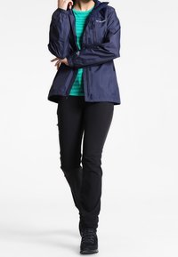 Marinblå jacka med huva, vattentåligt material, framfickor med dragkedja, bärs över en grönrandig skjorta, ihop med svarta byxor och stövlar.