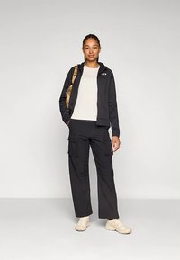 Black zip-up jacket over a white shirt, paired with black cargo pants and beige shoes. A brown backpack is slung over one shoulder.