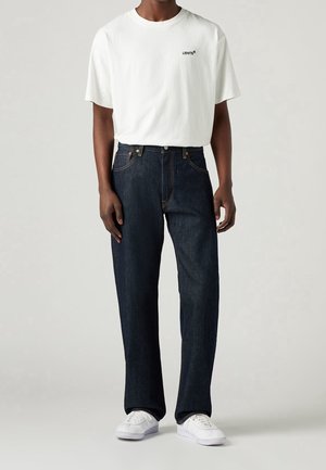 Mężczyzna ubrany w białą koszulkę Levi's, ciemnoniebieskie proste jeansy i białe sneakersy, stojący na tle jednolitego jasnego tła.