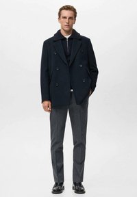 Cappotto doppiopetto blu navy in misto lana con colletto a scialle, abbinato a pantaloni grigi a fantasia e mocassini neri.