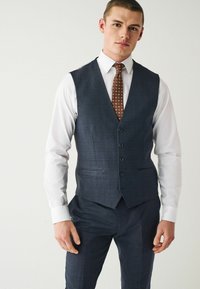 Gilet à carreaux bleu marine avec cinq boutons, revers crantés et poches latérales, associé à une chemise blanche et une cravate à motifs marron.