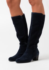 Knähöga boots i marinblå mocka med sidodragkedjor, klack i blockstil och en dekorativ knytning vid ankeln. Slät textur och skräddarsydd passform.