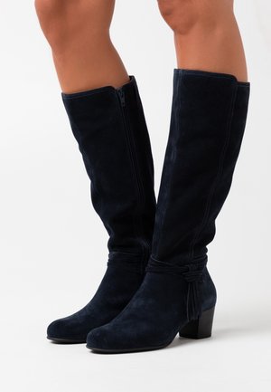 Bottes en daim bleu marine jusqu'aux genoux avec des fermetures éclair sur le côté, un talon bloc et un détail de lien décoratif à la cheville. Texture lisse et coupe ajustée.
