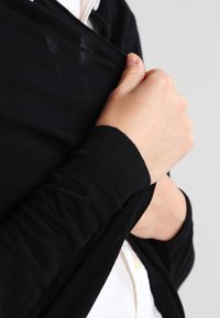 Černý, lehký pletený cardigan s dlouhými rukávy. Látka vypadá hladce; okraje jsou úhledně zpracované. Nositý přes bílou košili s límečkem.