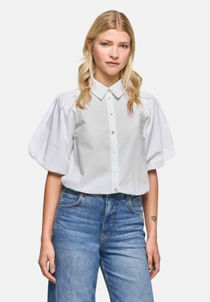 Woman with long blonde hair wears a white puff-sleeve button-up shirt and blue jeans, standing facing forward with a neutral expression.